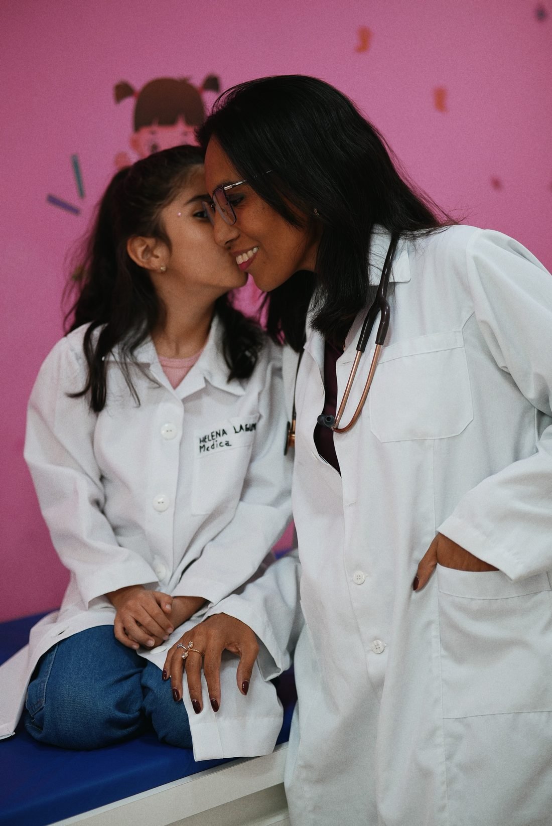 Dra. Vannia de jaleco recebendo um beijo no rosto da filha, que também veste um jalequinho escrito Helena Laguna, Médica
